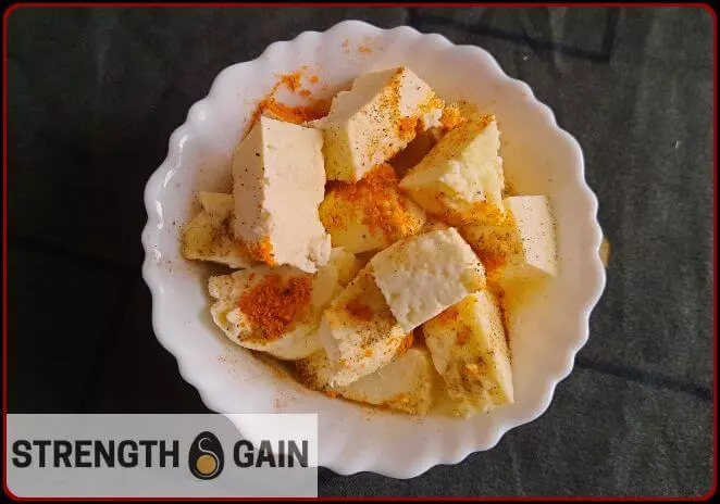 Cubes of raw paneer in a white bowl with salt pepper sprinkled on it.